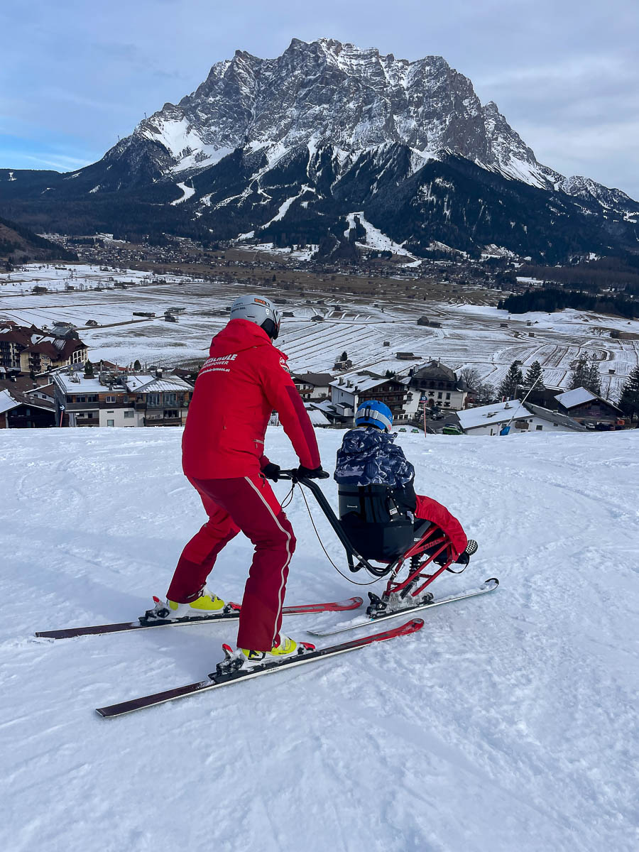 Skilehrer begleitet einen Gast im Bi-Ski bei einer Abfahrt mit Blick auf Lermoos und die Zugspitze in Tirol.
