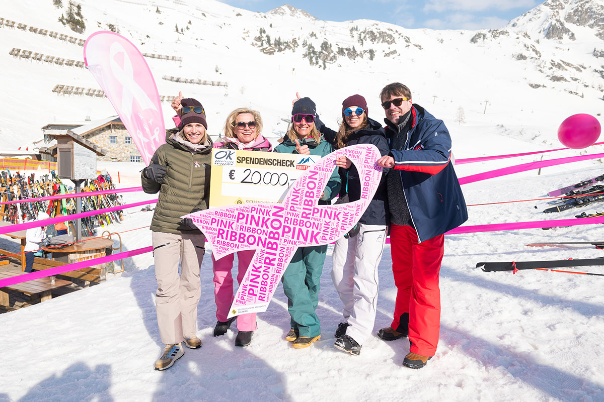 Fünf Personen im Skigebiet Obertauern präsentieren einen 20.000-Euro-Spendenscheck für Pink Ribbon und halten eine große pinke Schleife im Schnee.