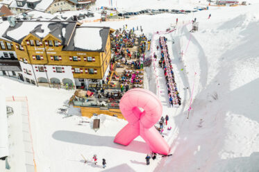 Große pinke Schleife aus Schnee vor einer Skihütte in Obertauern, daneben eine lange Tafel mit zahlreichen Teilnehmerinnen im Skigebiet.