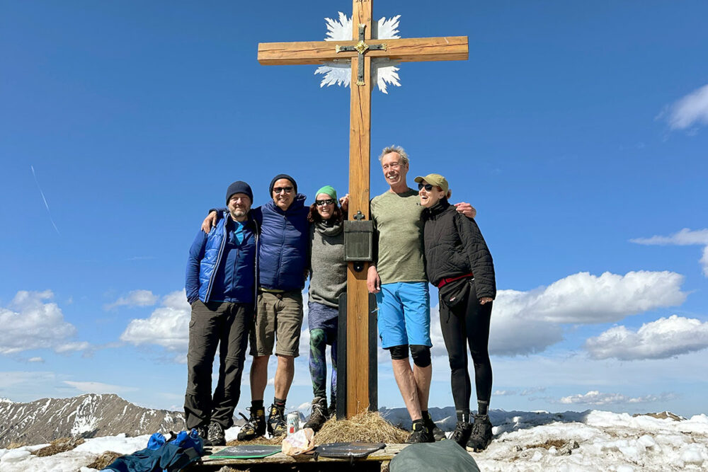 Wandergruppe am Gipfelkreuz des Hirschbichels in den Ammergauer Alpen im Frühling mit Schnee und blauem Himmel