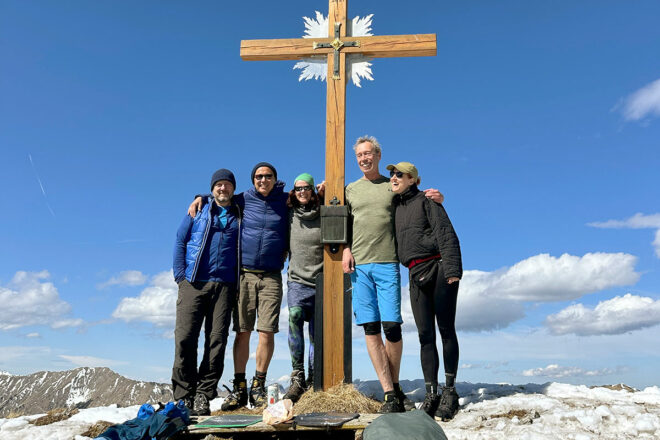 Wandergruppe am Gipfelkreuz des Hirschbichels in den Ammergauer Alpen im Frühling mit Schnee und blauem Himmel