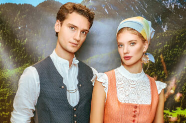 Modernes Trachtenpaar mit roséfarbenem Dirndl, Spitzenbluse und eleganter Herrenweste vor Alpenkulisse